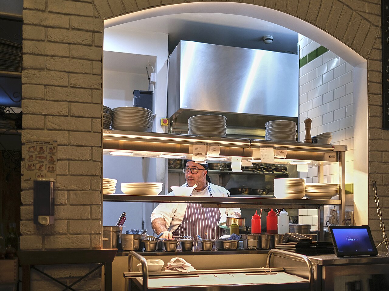 A chef working the pass during service, plates stacked on the shelf and order tickets clipped along the rail.