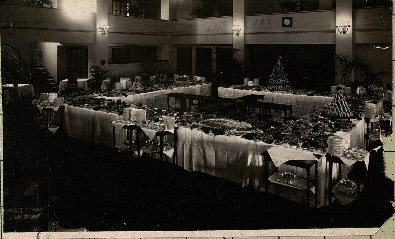 An archival photograph of a club dining room set for a banquet, with long tables of food and stacked plates.