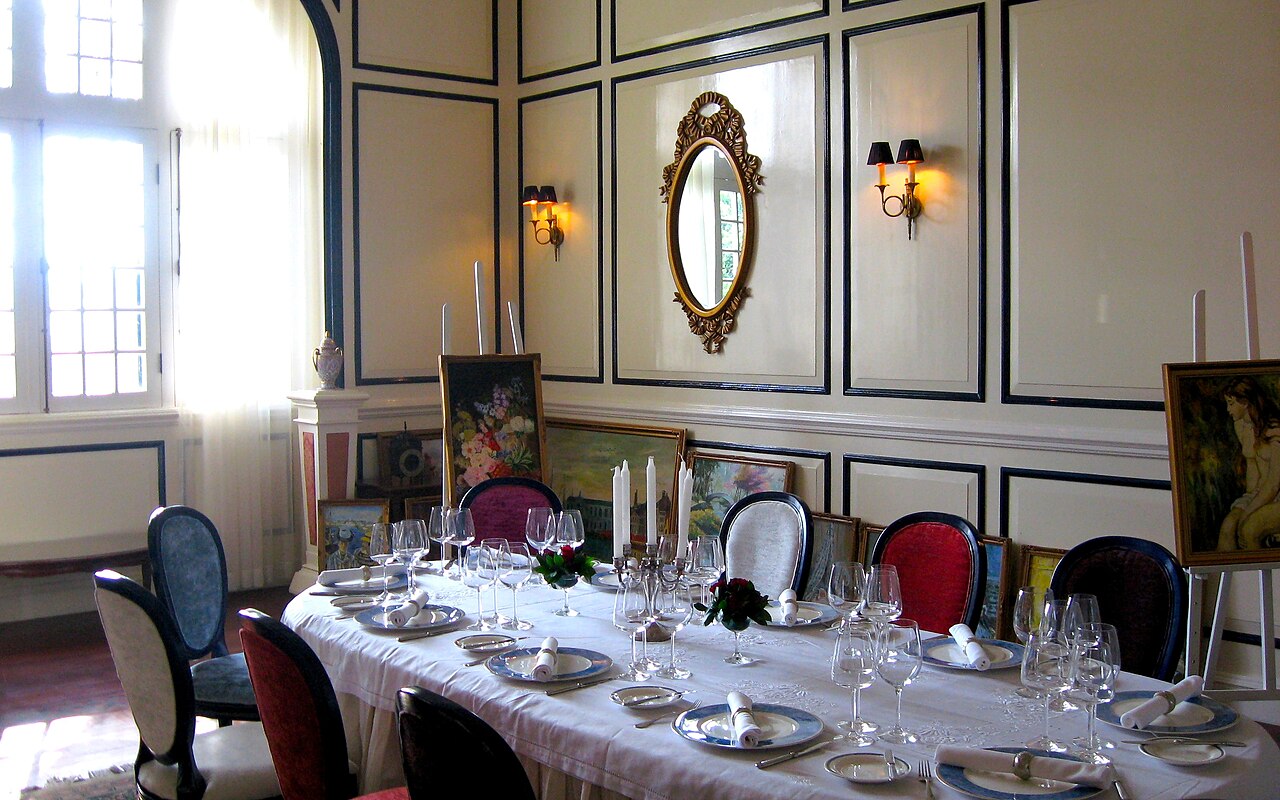 A small private dining room with an oval table laid for ten: white cloth, candles, crystal glasses and paintings on easels.