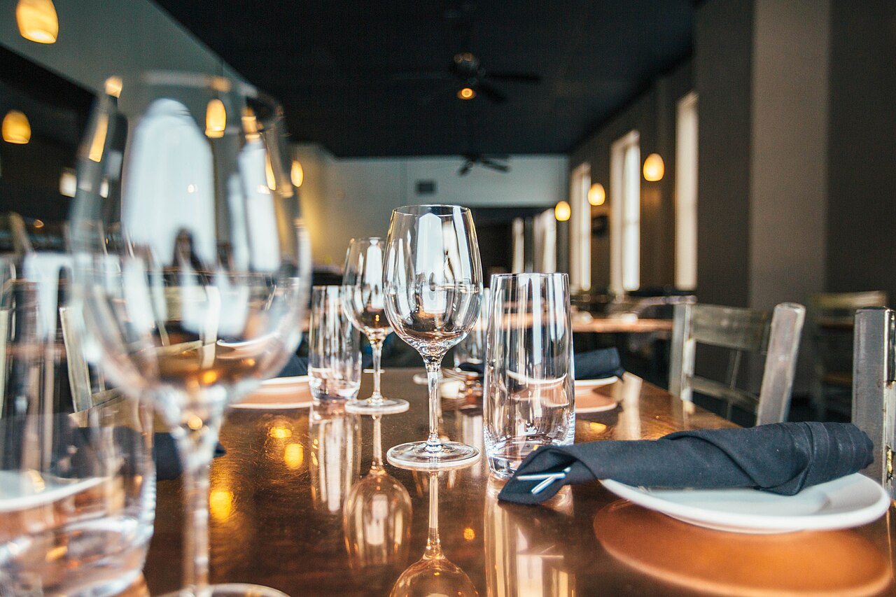 A restaurant table laid for dinner, with wine glasses, water glasses and a folded napkin on each plate, the dining room soft behind it.